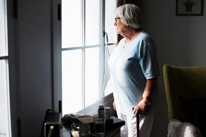 An older woman looks out the window deep in thought as she holds onto her walker